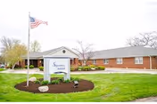 Front exterior of a single-story brick senior living facility with a landscaped lawn, entrance sign reading 'Signature HealthCARE at Parkwood', and an American flag.