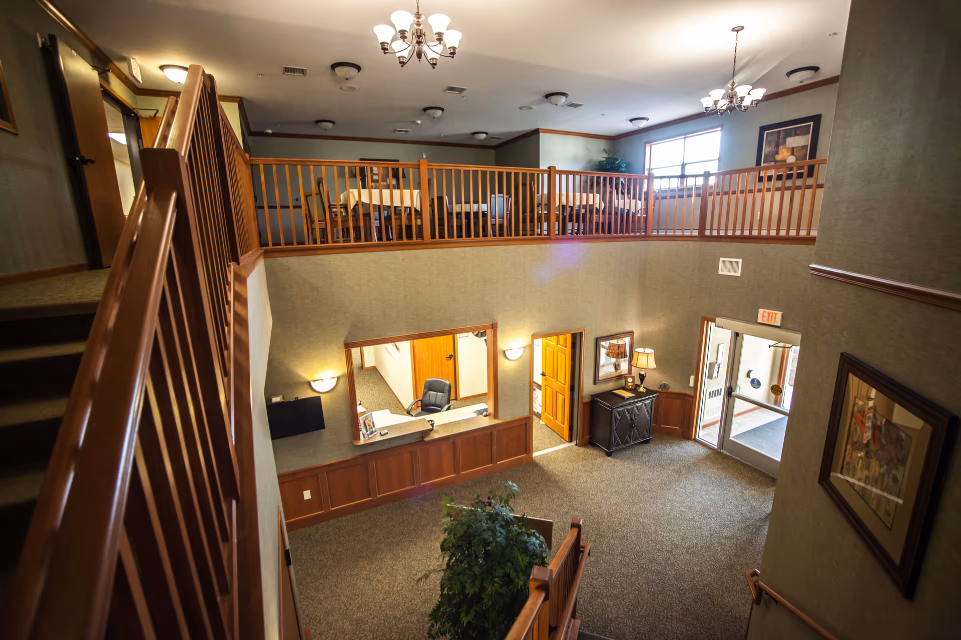 Interior view of a senior living facility showing a two-level common area with wooden railings, carpeted floors, and beige walls. The lower level has a reception desk with a chair, a small table with a lamp, framed pictures on the walls, and a glass door exit. The upper level has a dining area with tables and chairs, illuminated by ceiling lights and chandeliers.