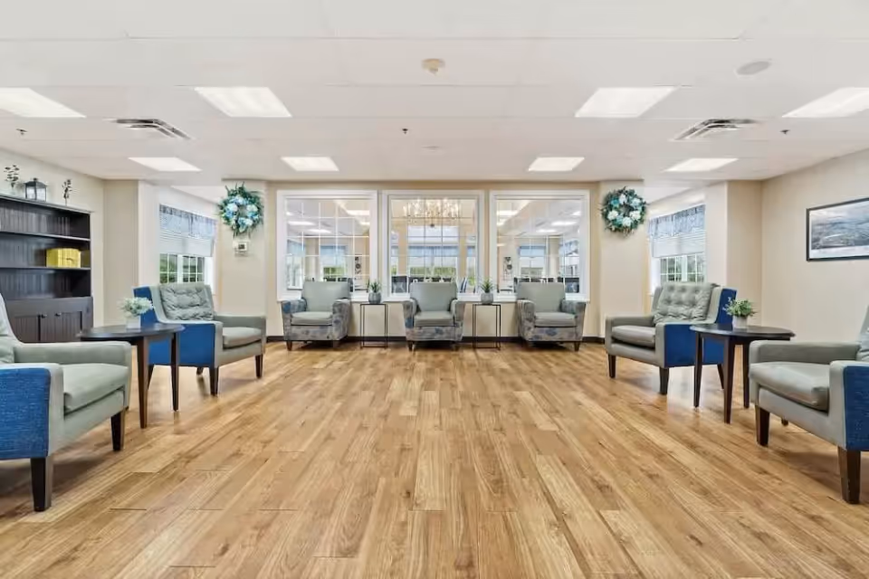 A spacious, well-lit common area with wooden flooring and multiple cushioned armchairs arranged along the walls. There are small side tables with plants next to some chairs, large windows letting in natural light, and decorative wreaths on the walls. A bookshelf is visible on the left side and a framed picture hangs on the right wall.