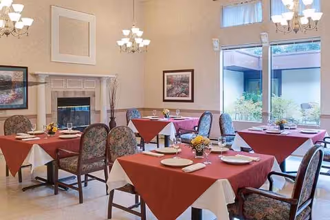 Senior living facility dining room with red-clothed tables, chairs, chandeliers, and large windows.