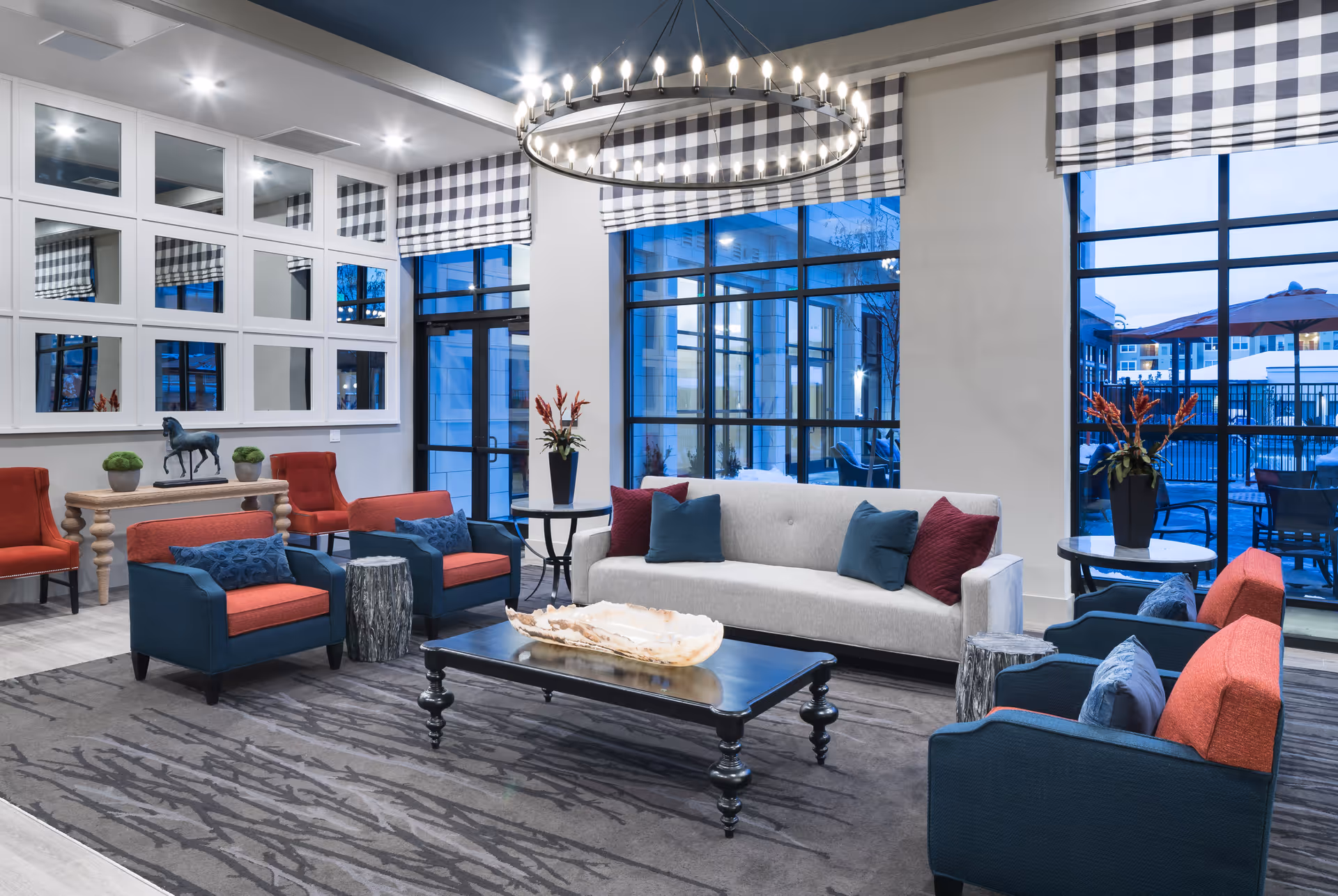A modern living room with a large gray sofa adorned with blue and red pillows, four armchairs with blue and red upholstery, a black coffee table with a decorative centerpiece, and a circular chandelier hanging from the ceiling. The room has large windows with black frames and checkered Roman shades, letting in natural light and showing an outdoor patio area with tables and umbrellas. There is a console table with decorative items and a wall of square mirrors reflecting the room.