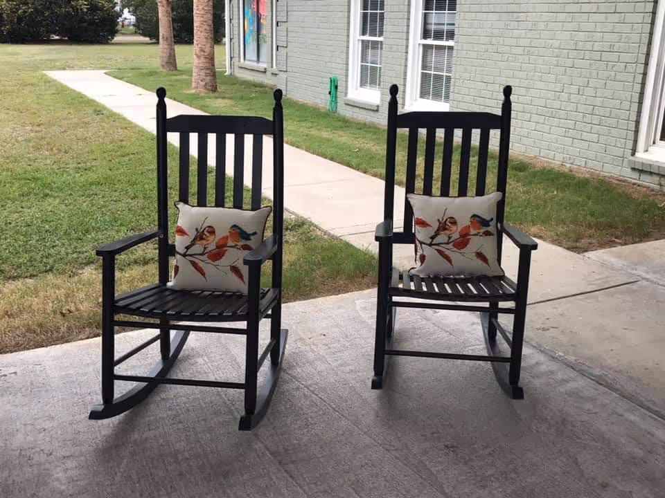 Two black wooden rocking chairs with cushions featuring bird designs are placed on a concrete patio outside a building with light green brick walls and white-framed windows. A grassy area and a sidewalk are visible in the background.
