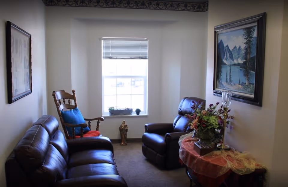 A cozy living room with a dark brown leather sofa and matching recliner chair. There is a wooden rocking chair with blue and red cushions near a window with blinds. A small table covered with a red and gold cloth holds a floral arrangement and a decorative lamp. Two framed paintings hang on the walls, one depicting a mountain lake scene and the other an abstract design.