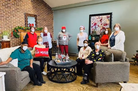 A group of nine people wearing face masks gathered in a living room with a brick fireplace, a decorative mirror, and a floral painting on the wall. Some are seated on sofas around a round coffee table with holiday decorations, and others are standing behind them.