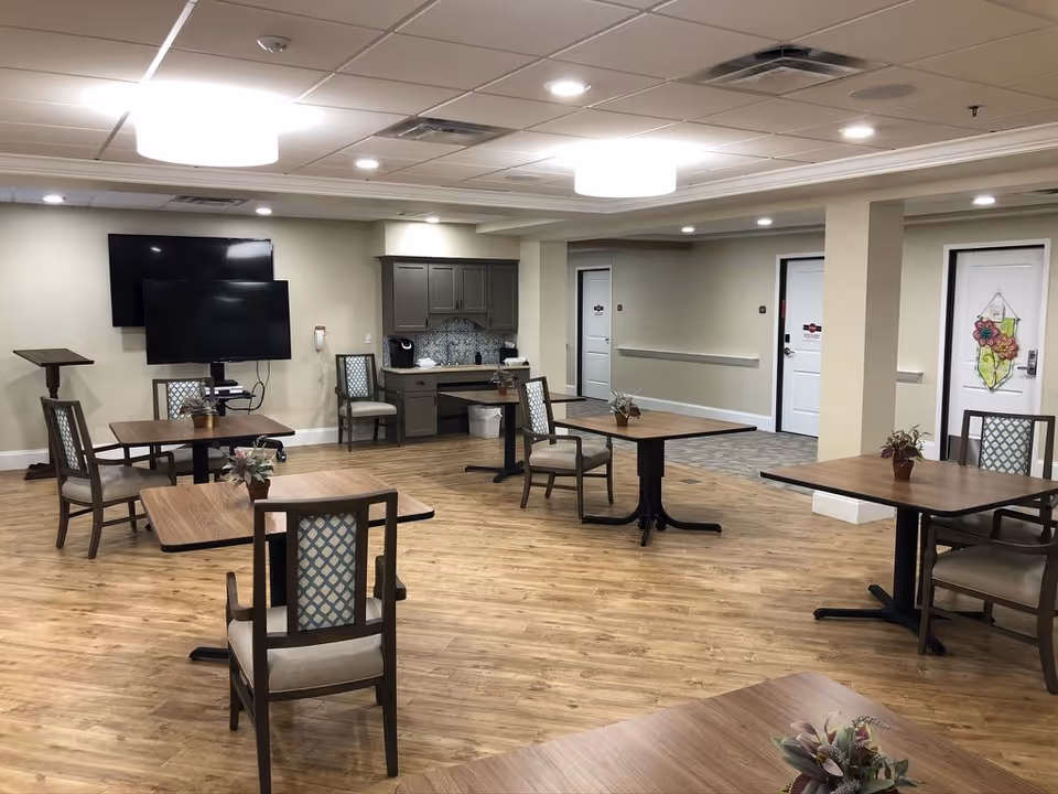 A spacious common area with wooden flooring, several tables each surrounded by chairs with patterned backs, and small potted plants on the tables. The room has a beige wall with a mounted TV and a smaller screen on a stand. There is a small kitchenette area with cabinets and a coffee maker. The ceiling has recessed lighting and two large ceiling lights. There are white doors along the back wall, one decorated with a floral hanging.