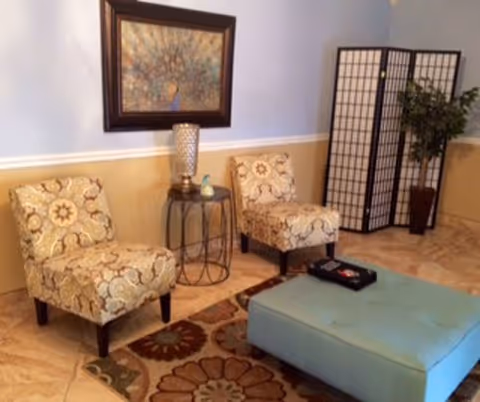 A cozy sitting area with two patterned armchairs, a small round side table with a decorative lamp and a small figurine, a large blue upholstered ottoman, a floral area rug, a folding room divider, a potted plant, and a framed artwork on the wall.