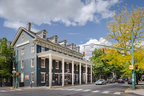 Street‑corner historic brick building with a white columned porch, traffic lights, cars, and trees under a partly cloudy sky.
