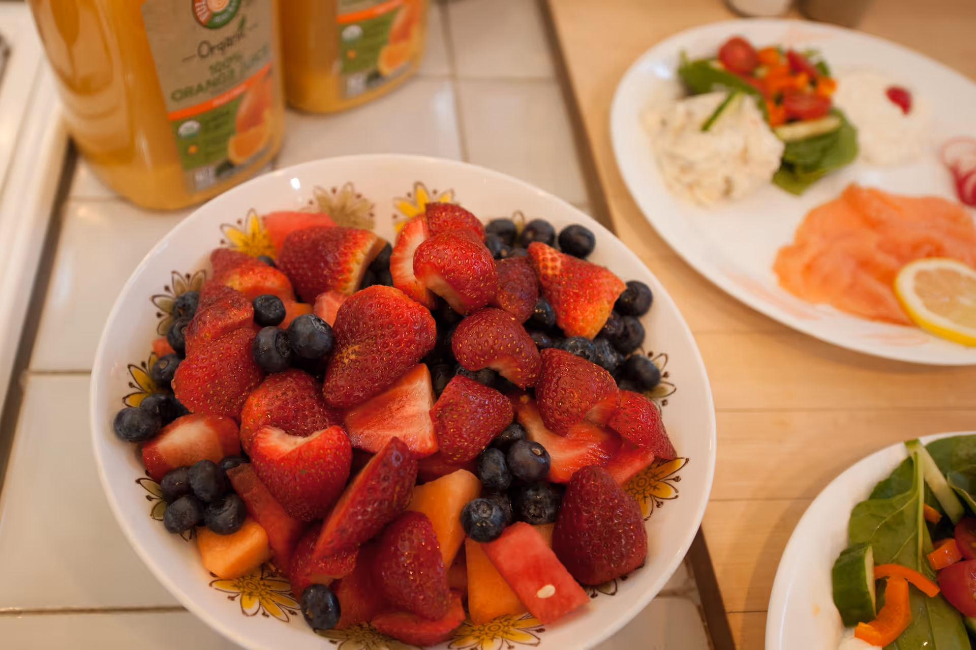 A bowl filled with fresh fruit including strawberries, blueberries, watermelon, and cantaloupe on a kitchen counter. In the background, there are two bottles of organic orange juice and two plates with various foods including smoked salmon, salad, and a lemon slice.