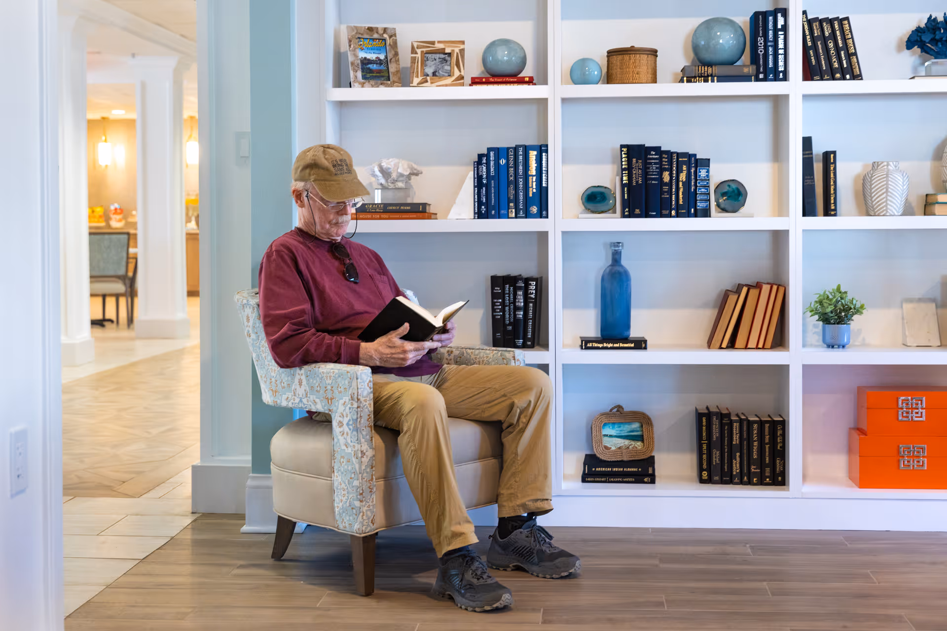 An elderly man wearing a cap, glasses, a maroon long-sleeve shirt, and khaki pants is sitting in a patterned armchair reading a book. Behind him is a white bookshelf filled with books, decorative items, and small plants. The setting appears to be a quiet, well-lit common area in a senior living facility.