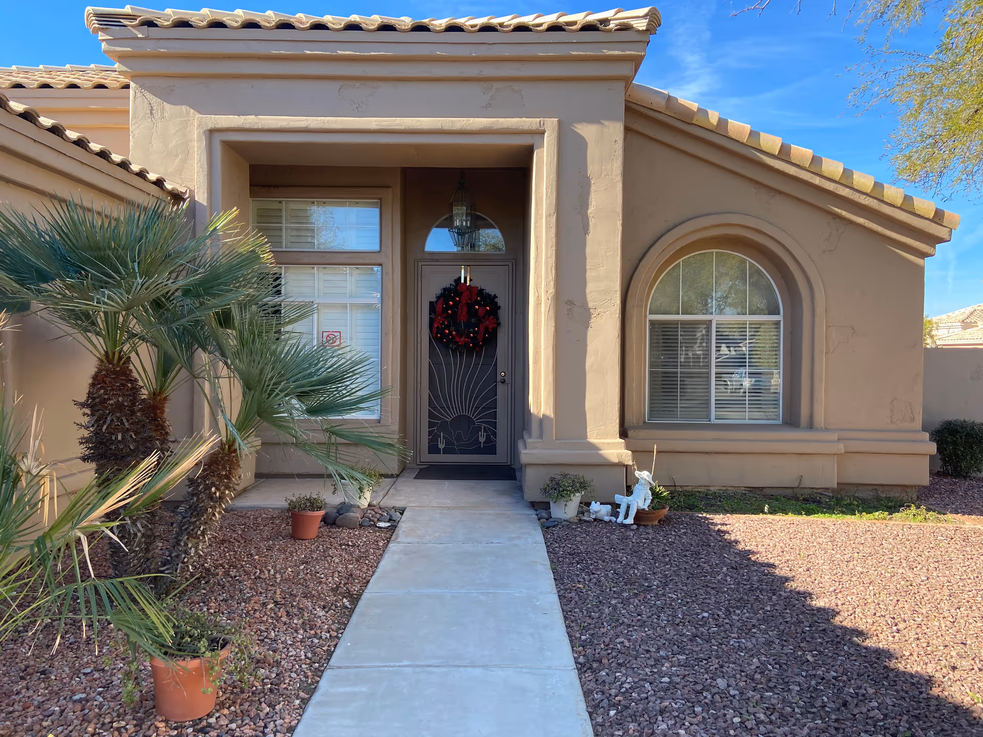 Front exterior view of a beige stucco house with a tiled roof, a walkway leading to a front door decorated with a red and green holiday wreath. There are two windows with white blinds on either side of the door, desert landscaping with small palm trees and potted plants, and a clear blue sky above.
