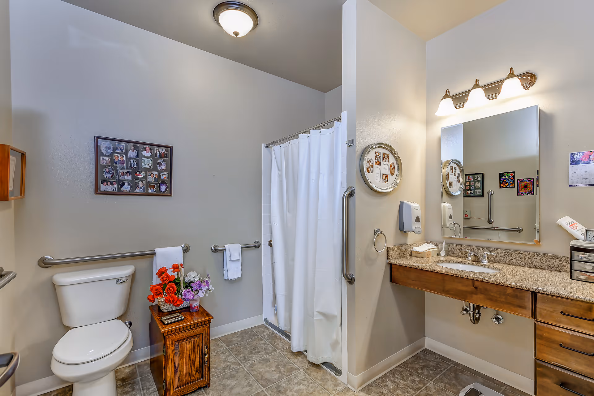 A clean and accessible bathroom with a toilet, a small wooden cabinet with flowers on top, a shower with a white curtain, a large mirror above a granite countertop with a sink, and grab bars installed on the walls for support.