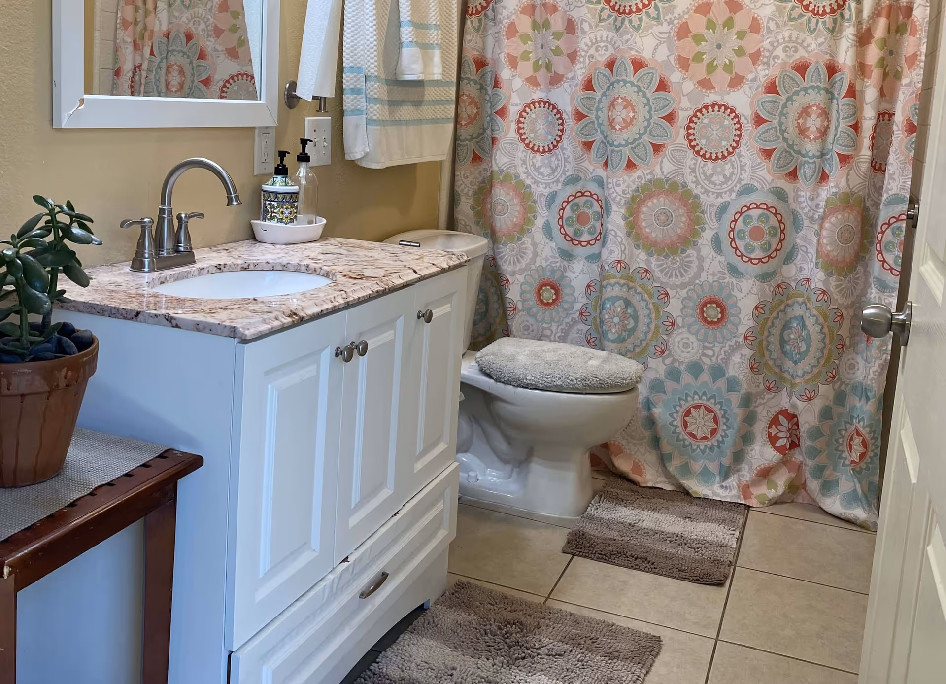 A bathroom with a white vanity cabinet topped with a marble countertop and a silver faucet. Above the vanity is a mirror and a towel hanging on the wall. Next to the vanity is a toilet with a soft gray cover. Behind the toilet is a colorful shower curtain with a floral pattern in shades of pink, blue, green, and white. The floor is tiled and has two gray bath mats. A small potted plant is on a wooden table beside the vanity.