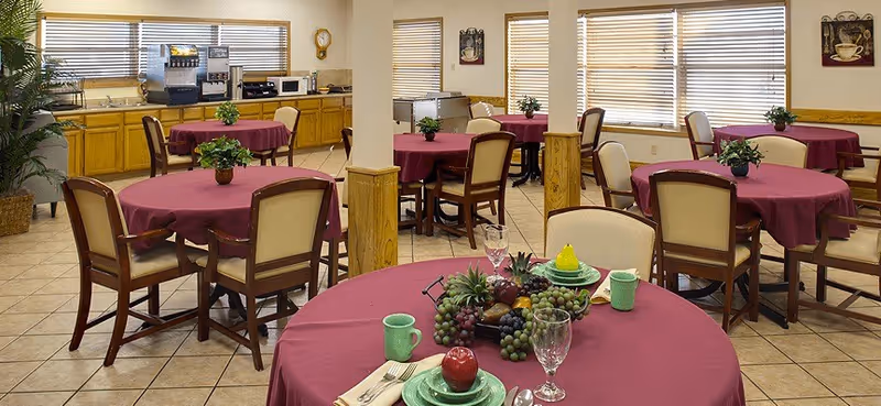 Dining area with multiple round tables covered with maroon tablecloths, each surrounded by wooden chairs with beige cushions. One table in the foreground is set with green plates, cups, silverware, a glass, and a centerpiece of assorted fruits. The room has tiled floors, large windows with blinds, and a counter along the back wall with coffee and beverage machines. Potted plants are placed on some tables and near the windows.