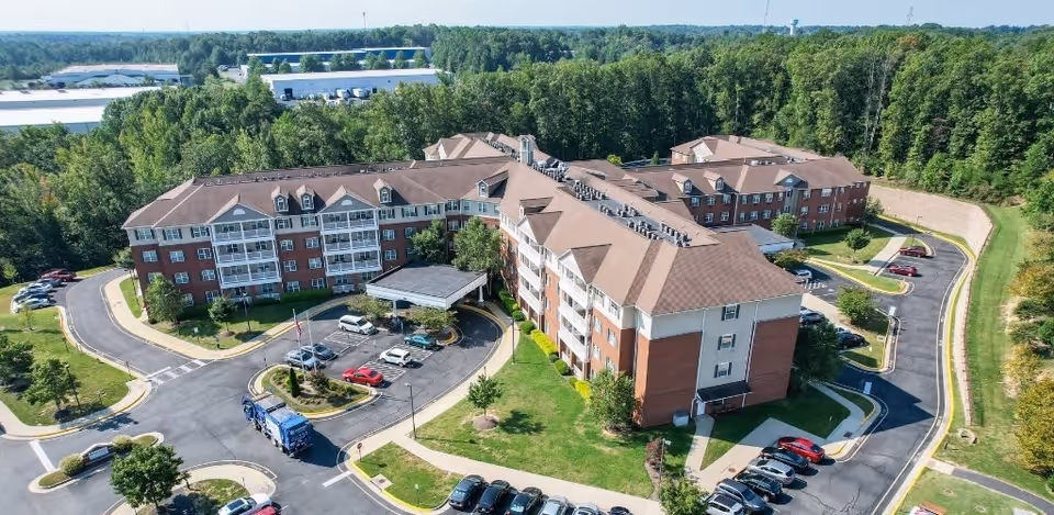 Aerial view of a large senior living facility named Harmony at Falls Run, featuring multiple connected brick buildings with brown roofs, surrounded by parking lots, roads, and green trees.