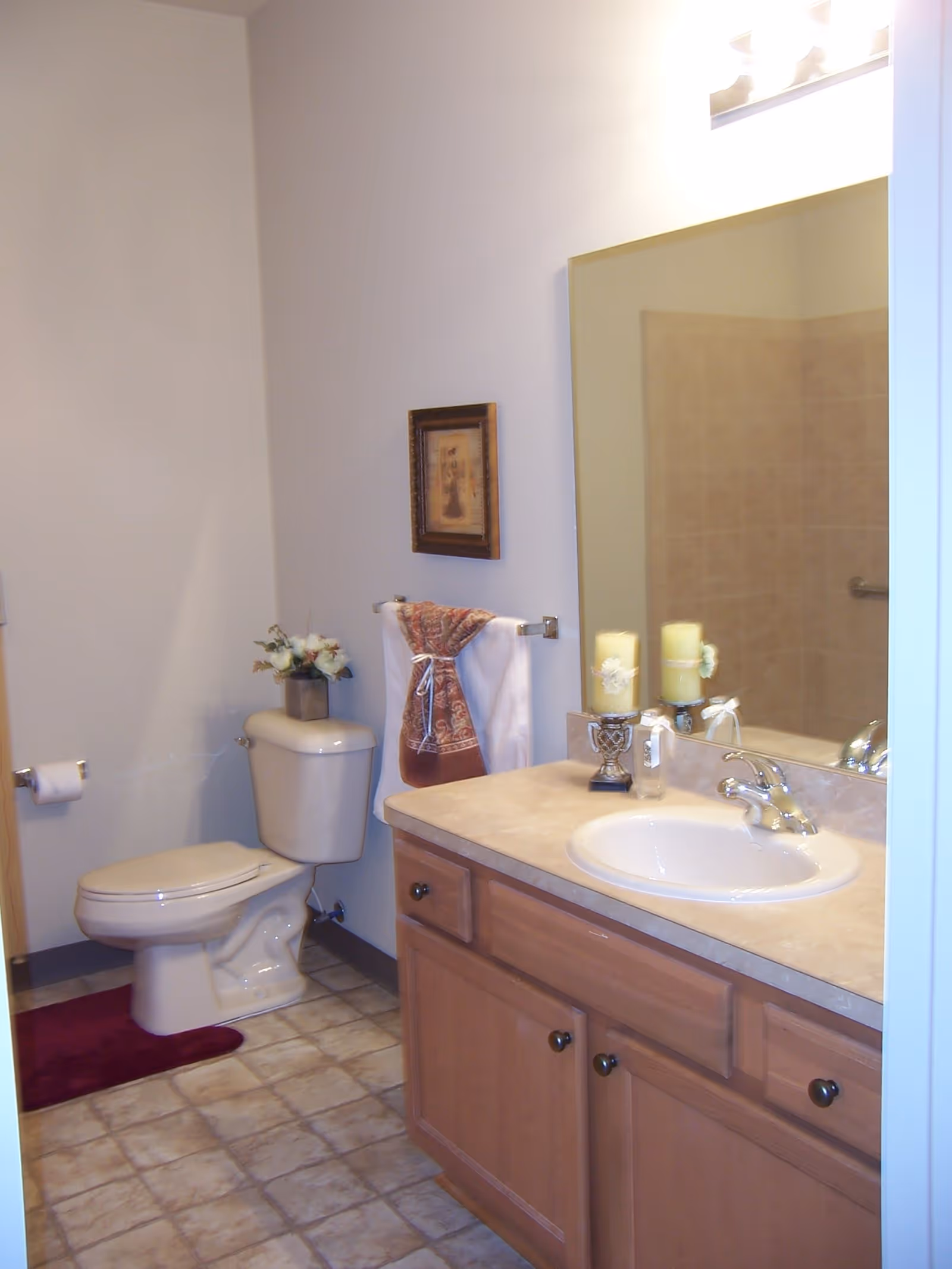 A clean bathroom with a toilet, a beige countertop with a sink, a large mirror above the sink, a towel rack with a decorative towel, a small framed picture on the wall, and decorative candles on the countertop.
