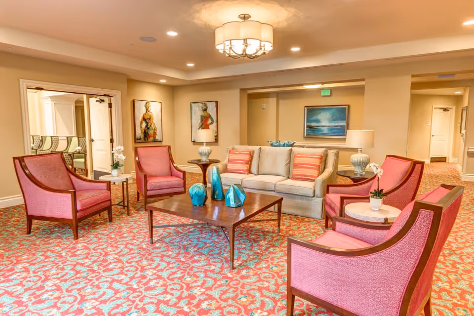 A cozy living room area with a beige sofa adorned with striped pillows, surrounded by four pink upholstered armchairs. A wooden coffee table with blue decorative vases sits in the center on a patterned red and blue carpet. The room is warmly lit by a ceiling light fixture and two table lamps on side tables. Artwork hangs on the beige walls, and a hallway is visible in the background.