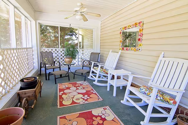 A screened-in porch with two white rocking chairs with floral cushions, two black metal chairs with a small round glass table between them, a ceiling fan, potted plants, and two colorful floral rugs on the floor. A decorative mirror with a flower frame hangs on the beige siding wall.