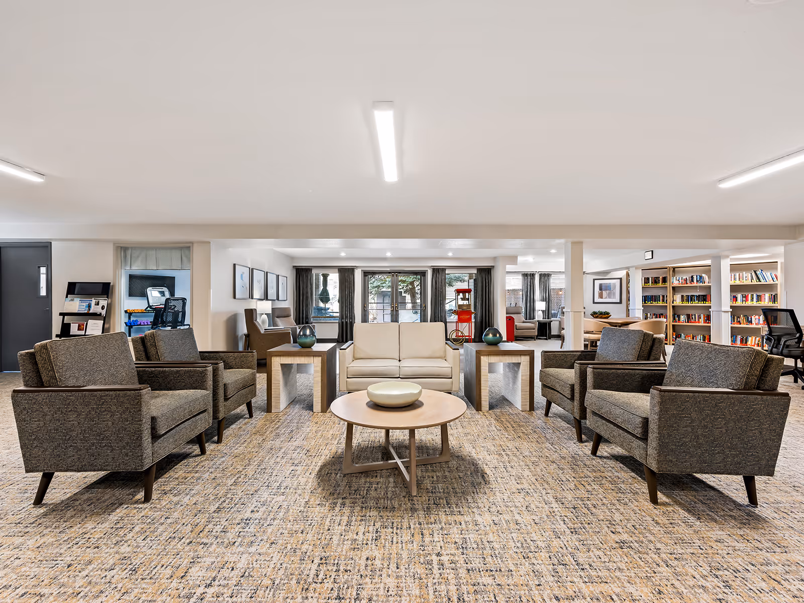 Spacious common lounge with sofas and armchairs arranged around a central coffee table, bookshelves and windows in the background.