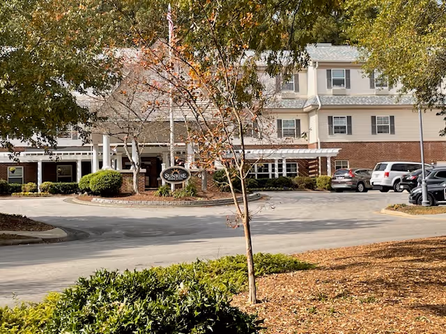 Front exterior of the Sunrise at North Hills senior living building with an entrance canopy, circular driveway, sign, and parked cars.