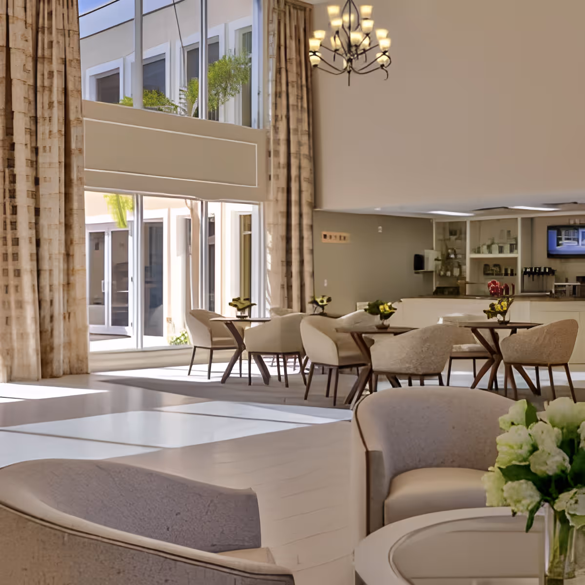 A bright and spacious common area with large windows allowing natural light to fill the room. The space features several round tables with beige upholstered chairs, small flower arrangements on each table, and a chandelier hanging from the ceiling. In the background, there is a counter area with shelves and a television mounted on the wall.