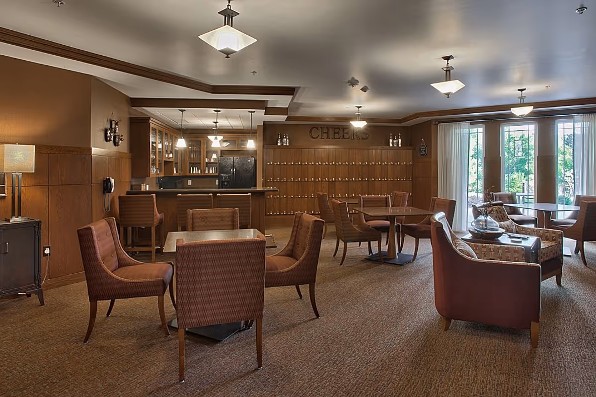 A cozy common area with multiple seating arrangements including armchairs and dining chairs around tables. The room features warm brown tones with wood paneling on the walls, a small bar area with bar stools, and large windows with sheer curtains letting in natural light. The word 'CHEERS' is displayed on the back wall above a series of cubby holes or lockers.