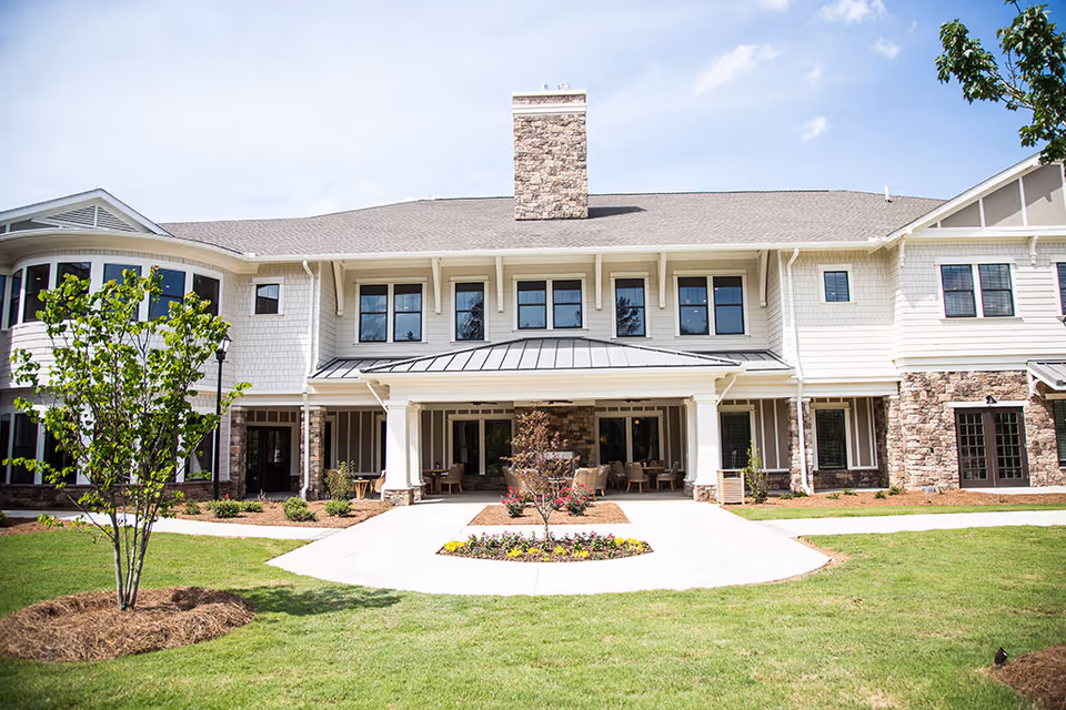 Exterior view of a two-story senior living facility building with a stone chimney, white siding, multiple windows, and a covered entrance with outdoor seating. The foreground features a well-maintained lawn, small trees, and a flower bed along a concrete walkway leading to the entrance.