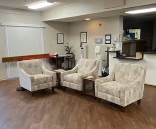 A seating area with three beige upholstered armchairs arranged around a small wooden table on a wooden floor. In the background, there is a counter with a TV mounted above it, some framed pictures on the wall, and a water dispenser. The room has neutral-colored walls and a window with vertical blinds.