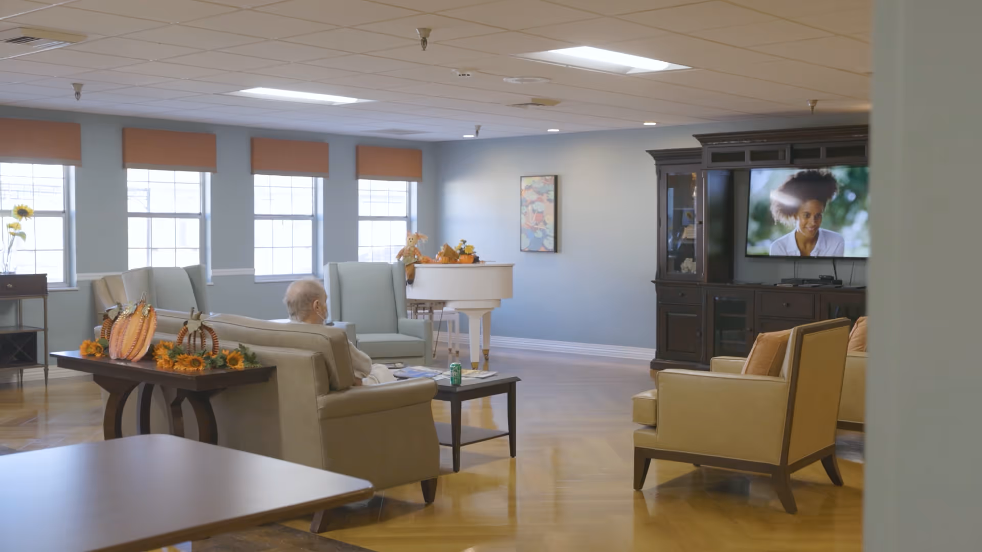 A senior living facility common area with comfortable seating including sofas and armchairs arranged around a coffee table. A person is seated on a sofa watching a television mounted on a dark wooden entertainment center. The room has large windows with orange blinds, a white piano in the corner, and decorative fall-themed items on a side table.