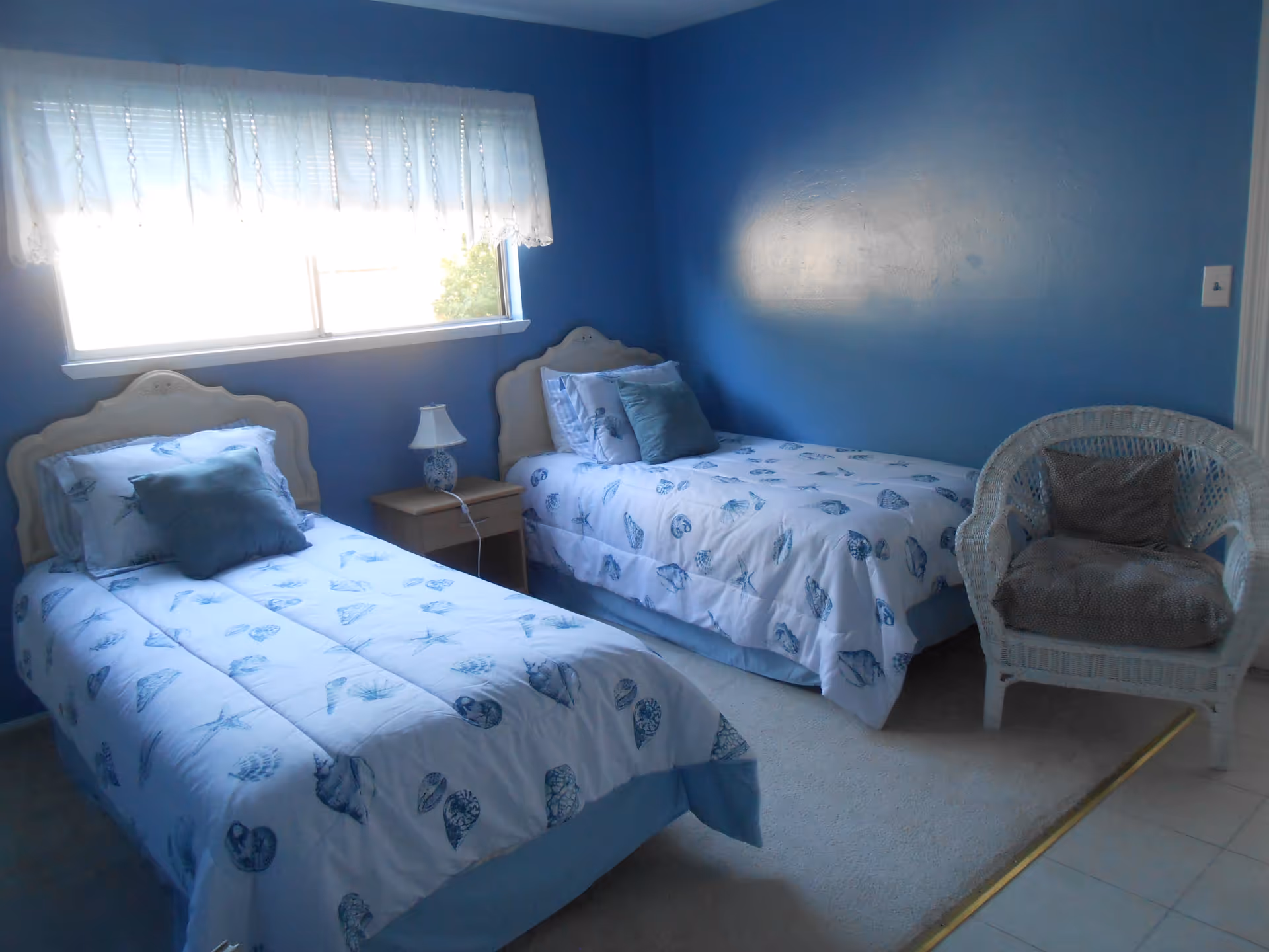 Small blue-painted bedroom with two twin beds, a nightstand and a wicker chair under a window.