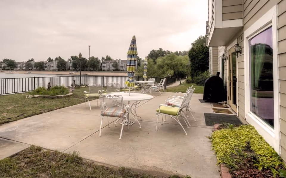 Outdoor patio area with white metal tables and chairs, some with striped cushions, and closed umbrellas. The patio overlooks a body of water with buildings on the opposite shore. There is a grill near the building entrance and greenery around the patio.