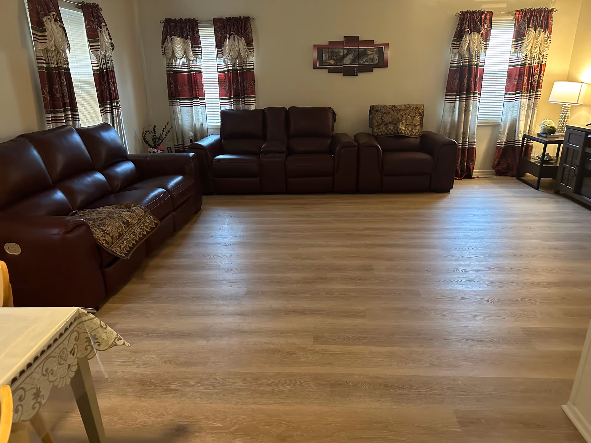 A living room with three brown leather sofas arranged against the walls, light wood flooring, three windows with red and white patterned curtains, a table with a lamp and decorative items on the right side, and a wall decoration above the middle sofa.