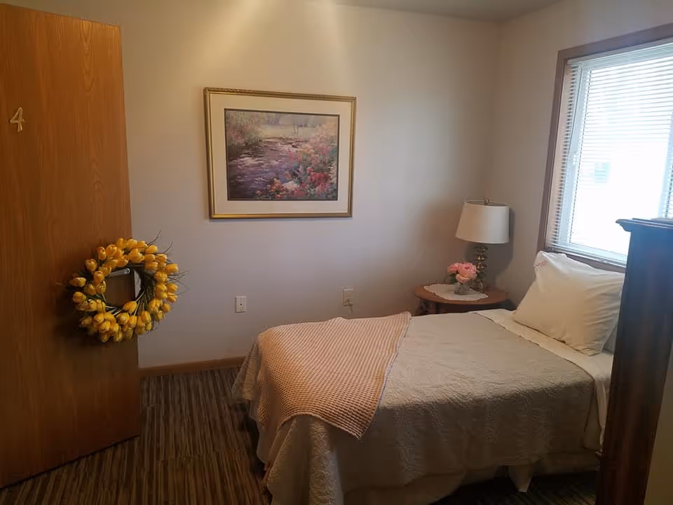 A small, cozy bedroom with a single bed covered in a beige quilt and a light pink throw blanket. There is a wooden door with the number 4 and a yellow flower wreath hanging on it. A framed painting of a nature scene hangs on the wall above the bed. Next to the bed is a wooden side table with a lamp and a small vase of pink flowers. A window with blinds allows natural light into the room.