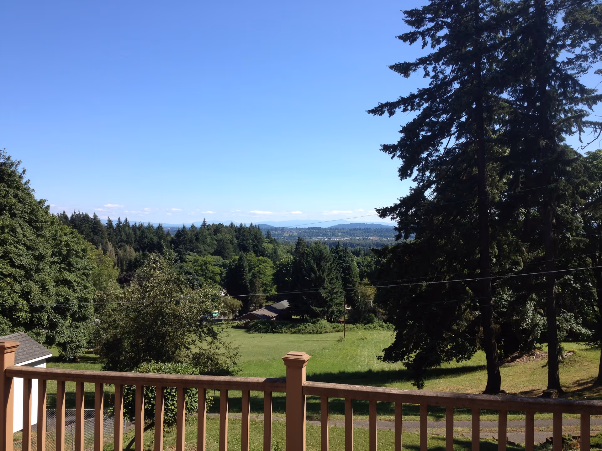 View from a wooden deck railing over a grassy yard and trees toward distant hills under a clear blue sky.