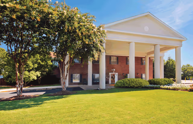 Front exterior view of a two-story brick building with large white columns supporting a triangular pediment at the entrance. The building is surrounded by green grass, bushes, and trees under a clear blue sky.