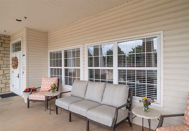 Covered front porch with cushioned seating, side tables with flowers, and a door with a wreath beside large windows.
