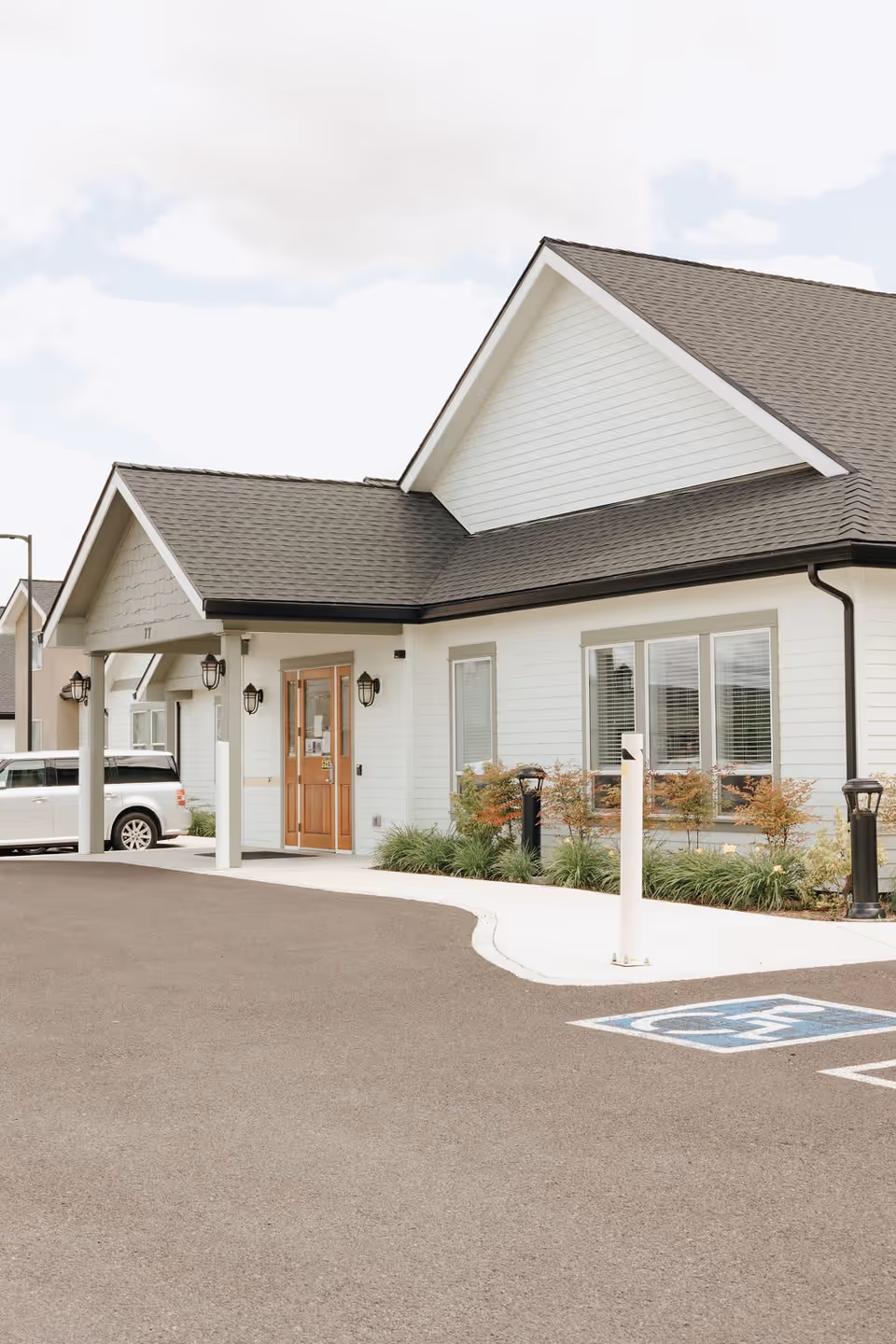 Exterior view of a single-story building with white siding and a dark shingled roof, featuring a covered entrance with wooden double doors. There are windows with blinds, landscaped bushes, and a handicapped parking space in front. A white vehicle is parked near the entrance.