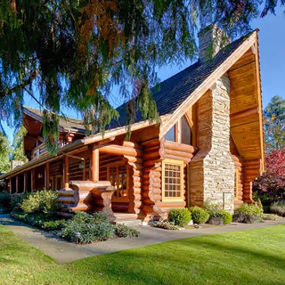 A large log cabin-style building with a steep roof and a prominent stone chimney, surrounded by green grass, shrubs, and trees under a clear blue sky.