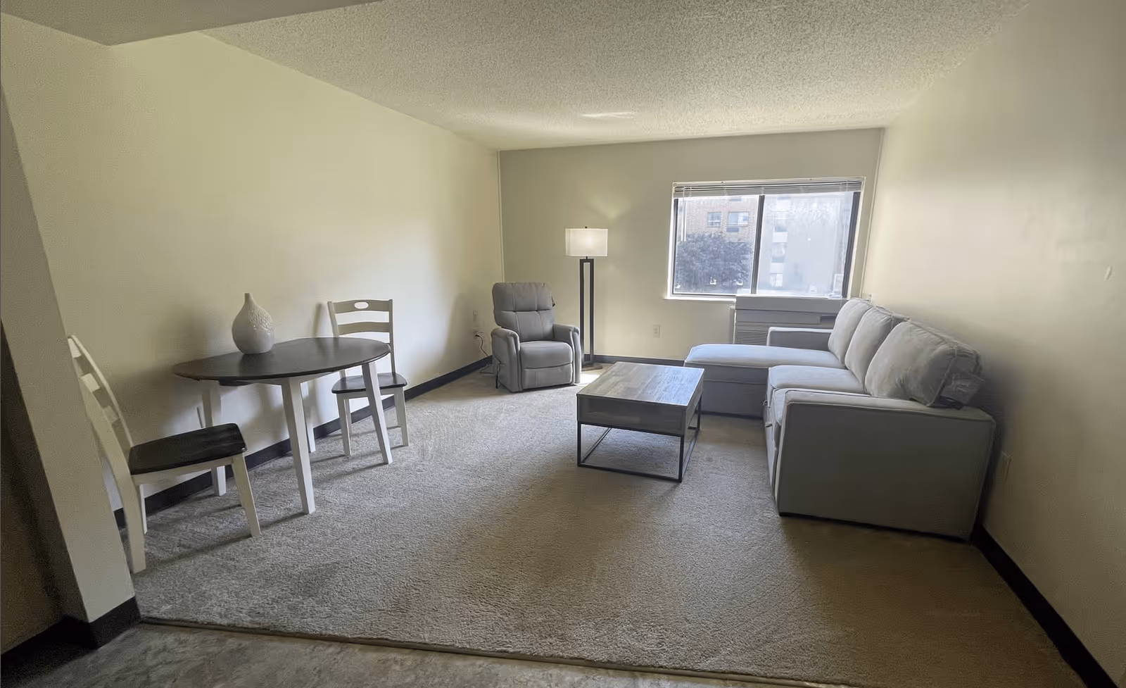 A senior living facility common area with a beige carpet, a gray sectional sofa, a gray recliner chair, a wooden coffee table, a small dining table with two chairs, a white decorative vase on the table, a floor lamp, and a window showing an outside view of buildings and trees.