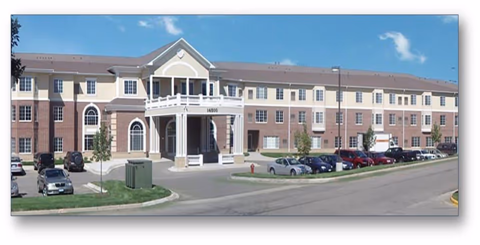 Exterior view of a large three-story senior living facility building with a covered entrance and multiple windows. Several cars are parked in the parking lot in front of the building under a blue sky with a few clouds.