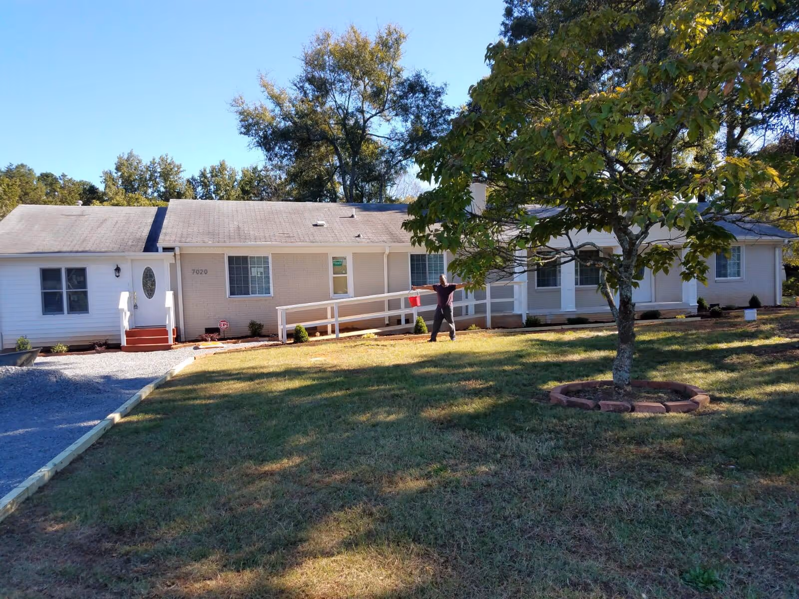 Single-story senior living facility exterior with a lawn, tree, and a person near the entrance.