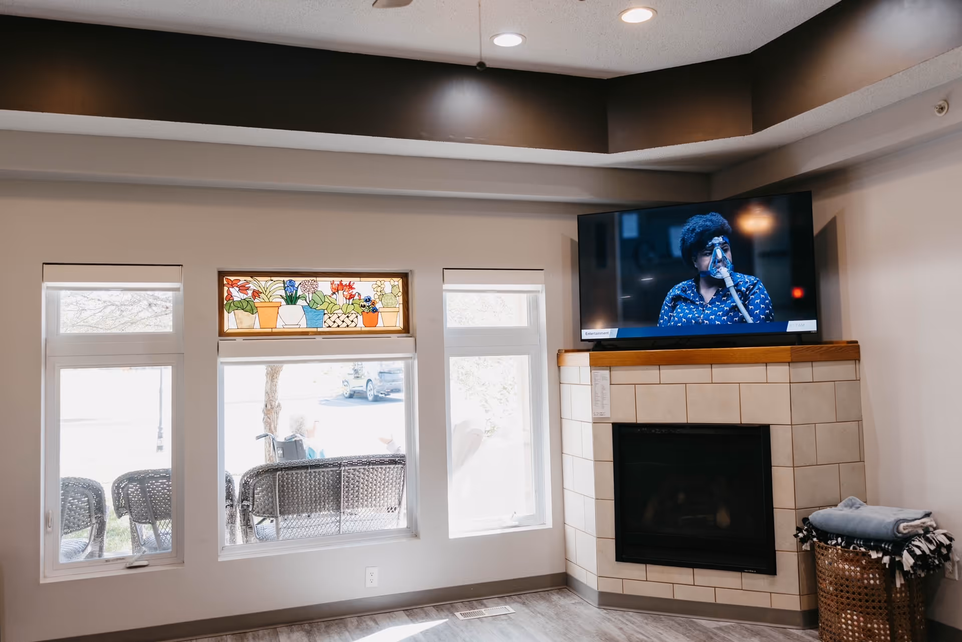 A cozy living room area with a tiled fireplace and a flat-screen TV mounted above it. The TV displays a person speaking into a microphone. There are three windows, one with a colorful stained glass panel featuring potted plants. Outside the windows, there are wicker chairs and a person sitting in a wheelchair. A basket with folded blankets is placed next to the fireplace.