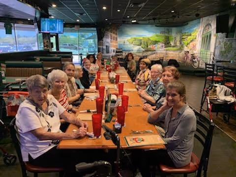 Elderly residents sitting around a long table in a dining room with murals on the walls.