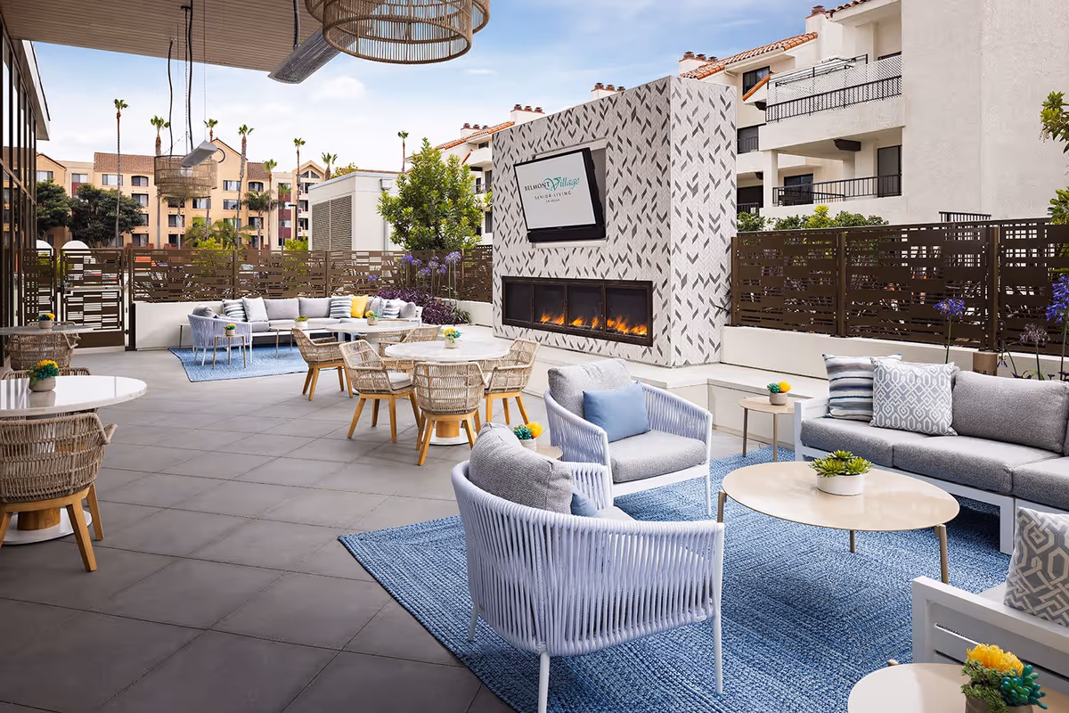 Outdoor patio area at Belmont Village Senior Living La Jolla featuring modern wicker and cushioned seating, round tables, a blue rug, a decorative fireplace with a mounted TV displaying the facility's logo, and surrounding buildings with palm trees in the background.