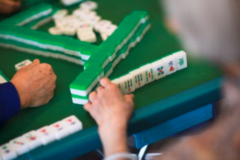 Close-up of two people playing a game of Mahjong on a green table, showing their hands and Mahjong tiles arranged in front of them.