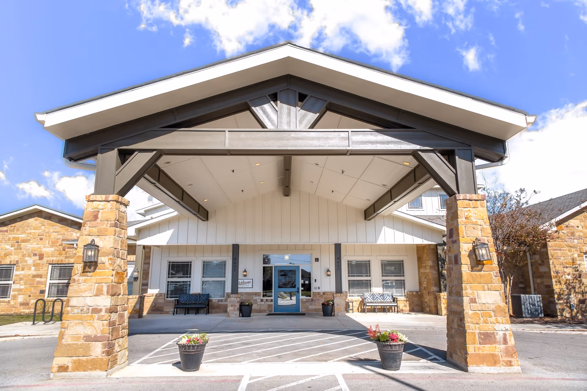 Front entrance of a senior living facility with a covered driveway supported by stone pillars, benches on either side of the entrance, potted plants, and a blue door under a clear blue sky with some clouds.