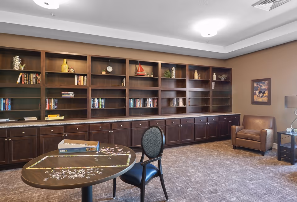 A quiet room with a large wooden bookshelf filled with books and decorative items, a round table with a partially completed puzzle and a box on it, a single chair, a brown leather armchair, a side table with a lamp, and a framed picture on the wall.