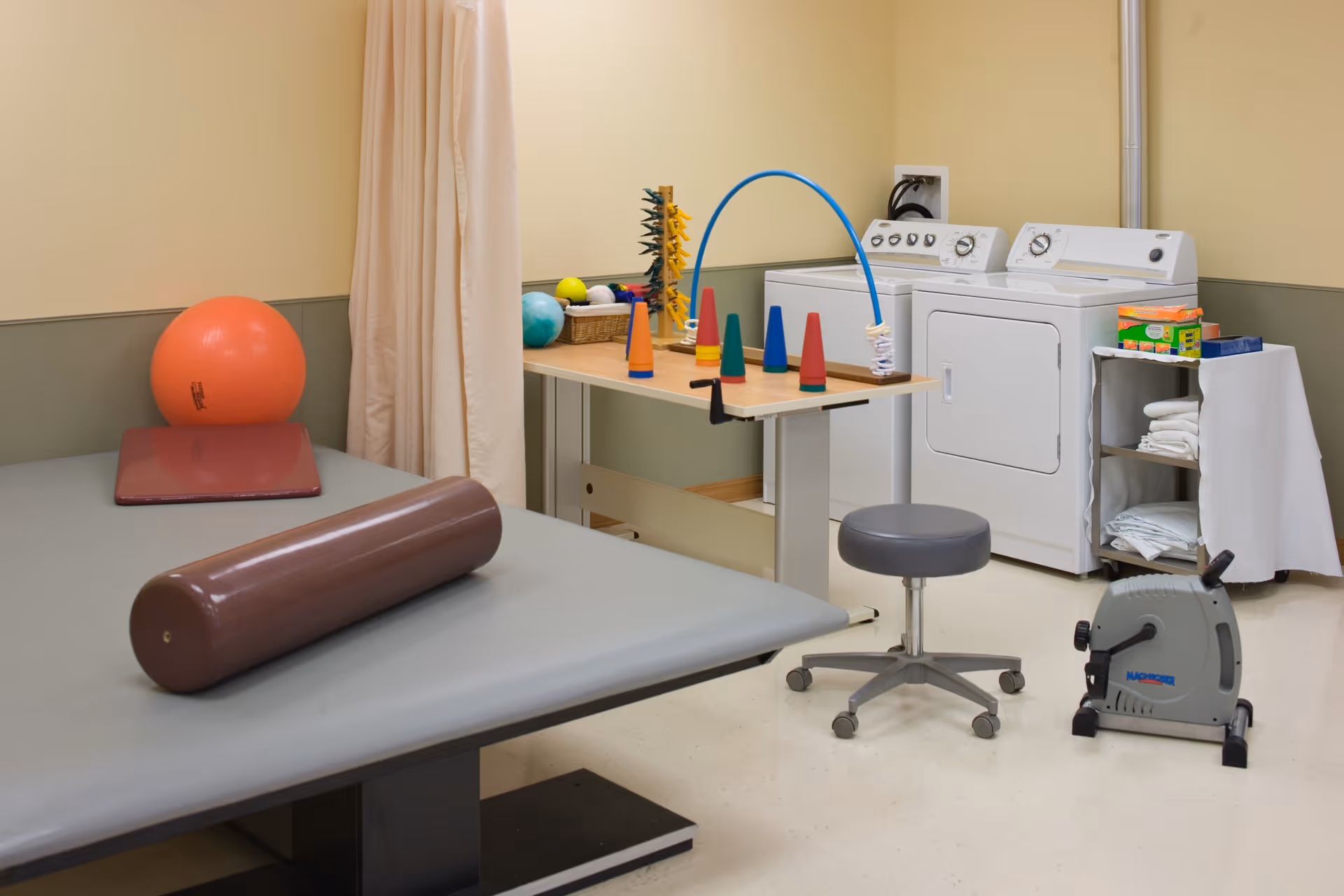 A therapy or rehabilitation room with a padded treatment table holding a brown cylindrical bolster, a red mat, and an orange exercise ball. Nearby is a table with colorful cones and therapy tools, a gray rolling stool, a small exercise bike, and a washer and dryer in the corner. The walls are beige with a green lower half, and there is a curtain divider.