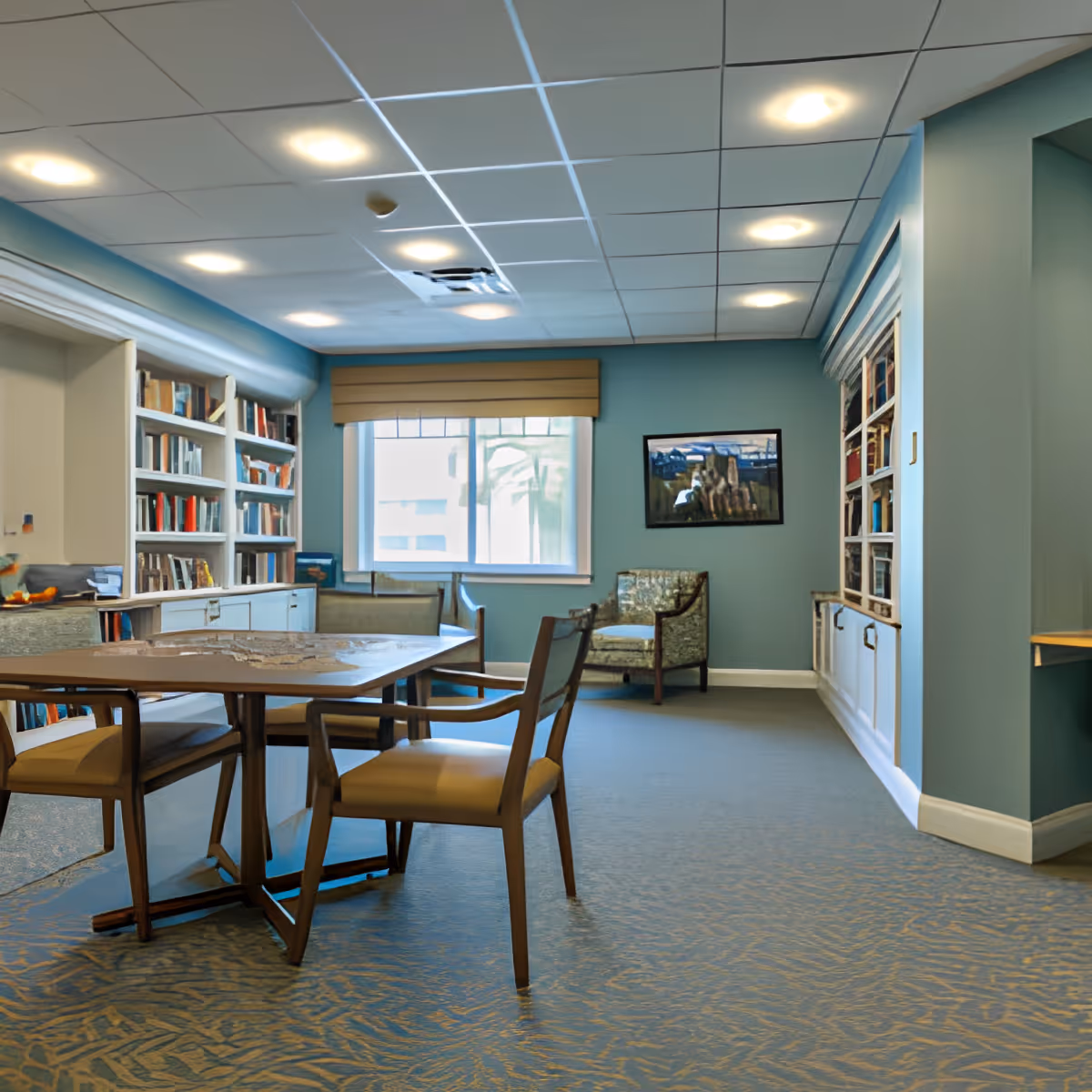 A cozy reading or activity room with a large wooden table surrounded by chairs, built-in bookshelves filled with books, a window with a beige blind, a framed painting on the blue wall, and an upholstered armchair in the corner.