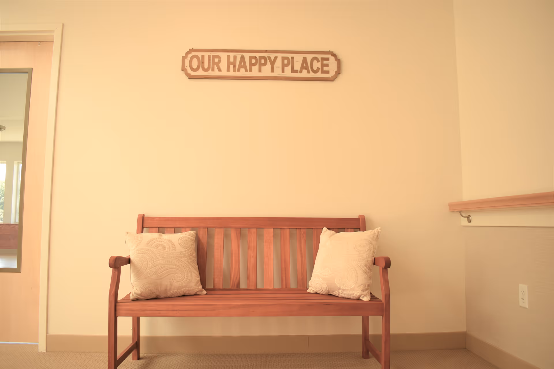 A wooden bench with two beige patterned cushions is placed against a light-colored wall. Above the bench, a wooden sign reads 'OUR HAPPY PLACE'. To the left, there is a door with a window panel, and to the right, a handrail is mounted on the wall.