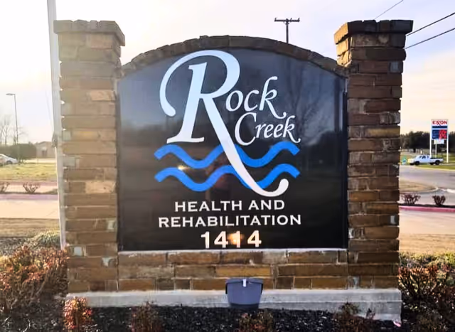 A stone-pillared roadside sign reading "Rock Creek Health and Rehabilitation 1414" with a blue wave logo.